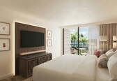 A hotel room with a large bed, a television mounted on the wall, a small table with chairs, and a balcony with a view of palm trees.