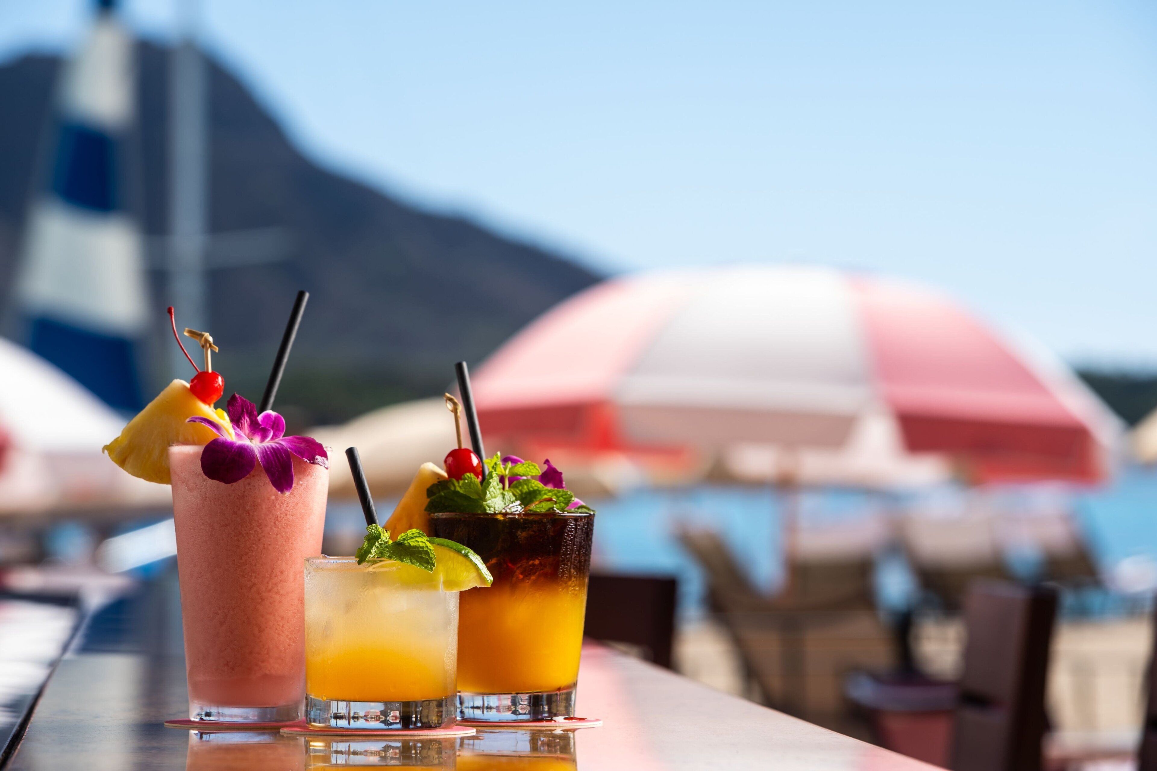 Three cocktails on a table with a mountain and umbrellas in the background.