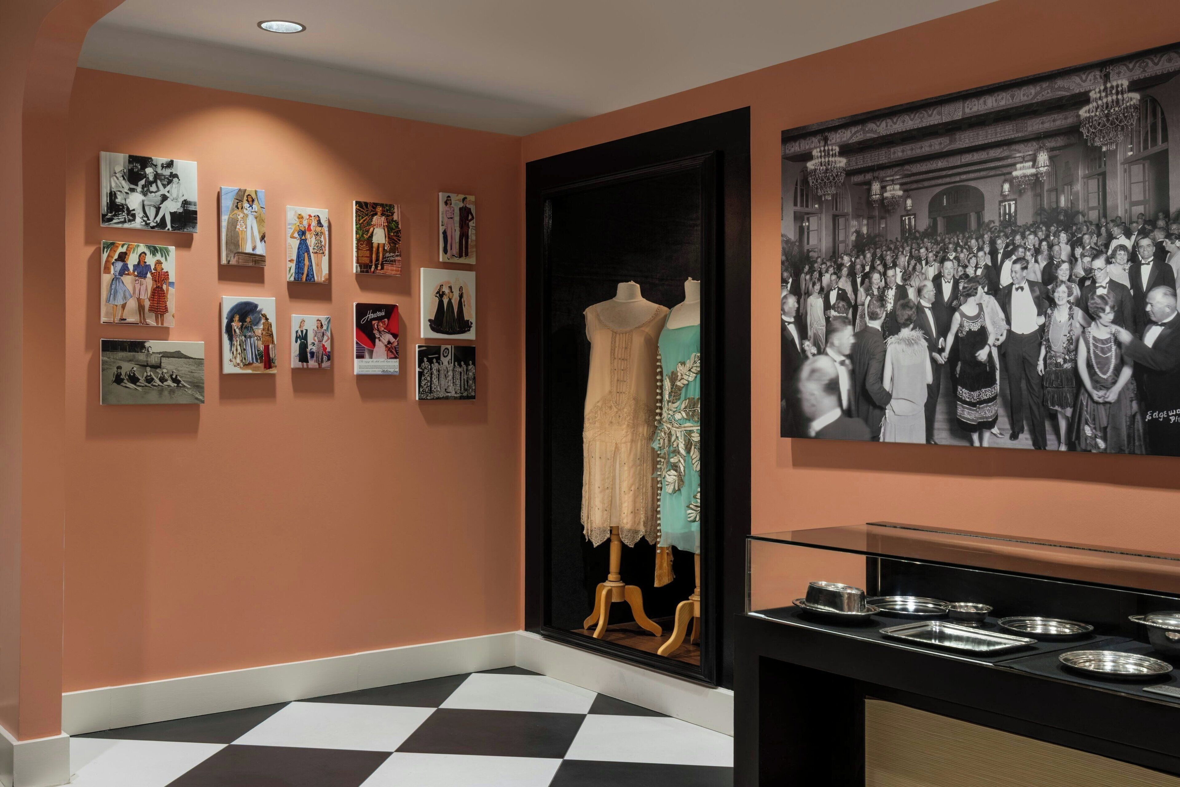 A hallway with a black and white checkered floor, a wall with framed photographs, and a mannequin in a dress.