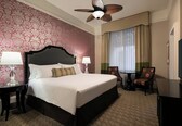 A hotel room with a large bed, two bedside tables with lamps, a ceiling fan, a TV, a desk, and a chair.