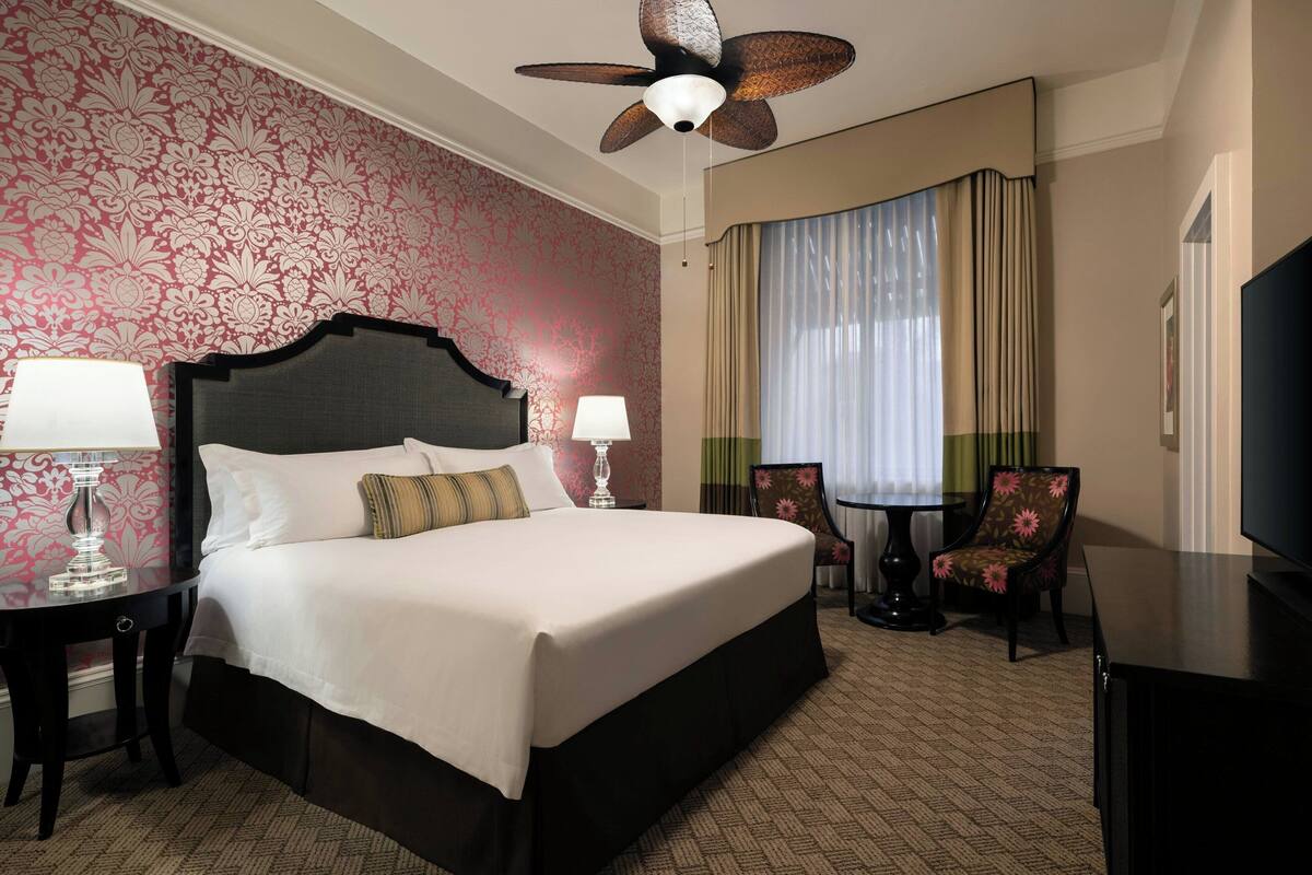 A hotel room with a large bed, two bedside tables with lamps, a ceiling fan, a TV, a desk, and a chair.