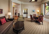 A hotel room with a sofa, armchair, dining table, and chairs. There is a ceiling fan, a lamp, and a window with curtains.