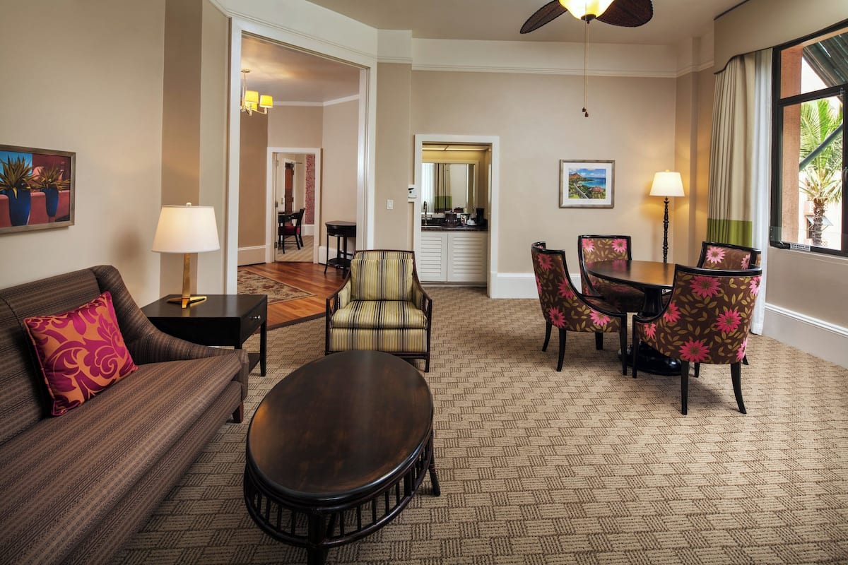 A hotel room with a sofa, armchair, dining table, and chairs. There is a ceiling fan, a lamp, and a window with curtains.