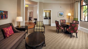 A hotel room with a sofa, armchair, dining table, and chairs. There is a ceiling fan, a lamp, and a window with curtains.