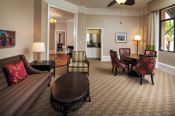 A hotel room with a sofa, armchair, dining table, and chairs. There is a ceiling fan, a lamp, and a window with curtains.