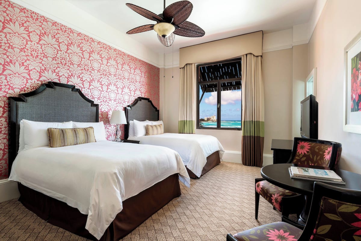 A hotel room with two beds, a ceiling fan, a view of the ocean, and a dining area with a table and chairs.