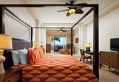 A four-poster bed with a patterned bedspread, a television, a desk with a chair, and a view of the ocean through a large window.