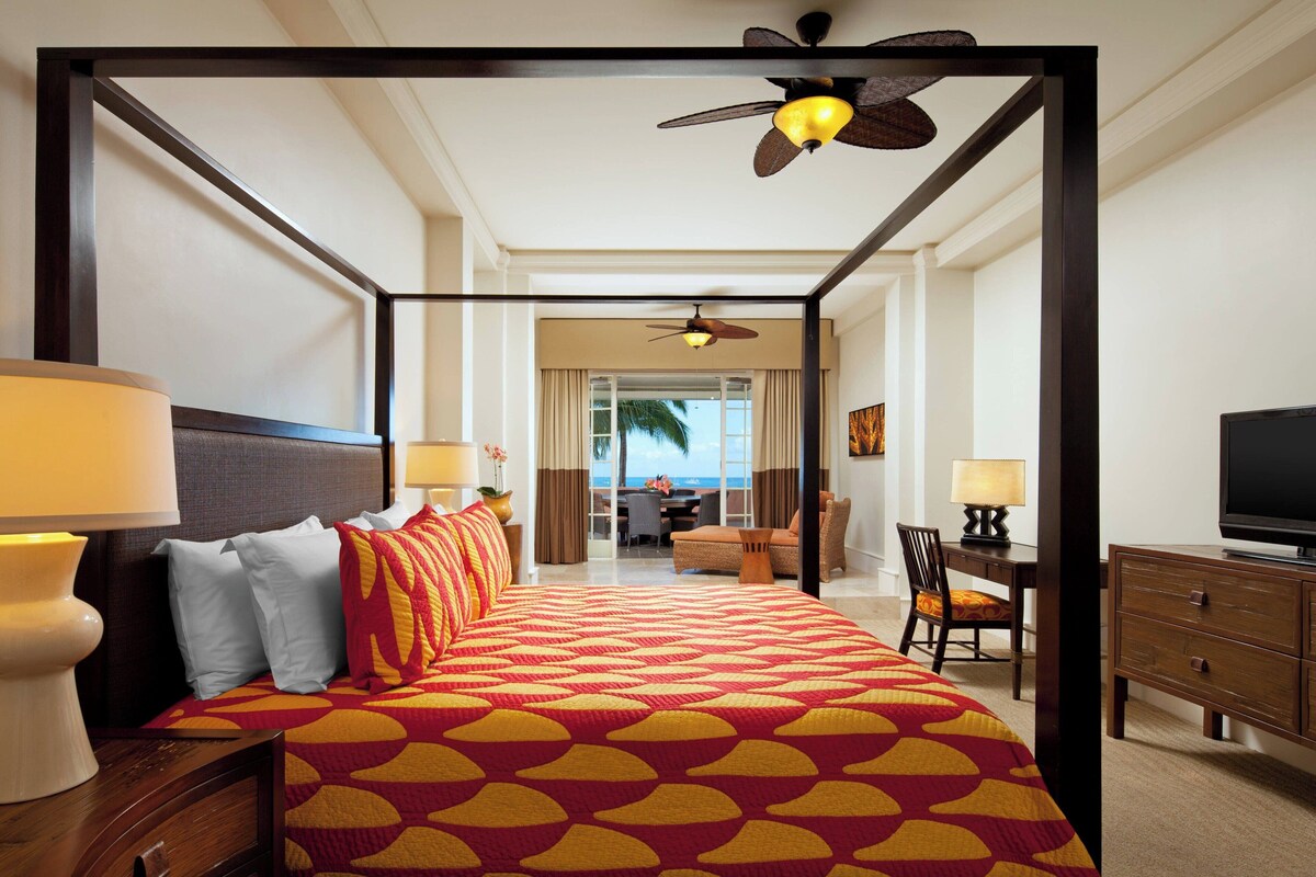 A four-poster bed with a patterned bedspread, a television, a desk with a chair, and a view of the ocean through a large window.