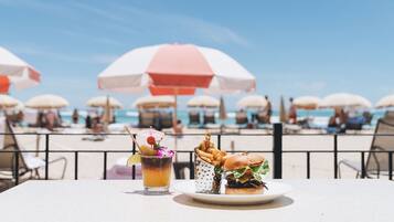 Een eettafel aan het strand met een hamburger, frietjes en een drankje, op de achtergrond ligbedden en mensen.