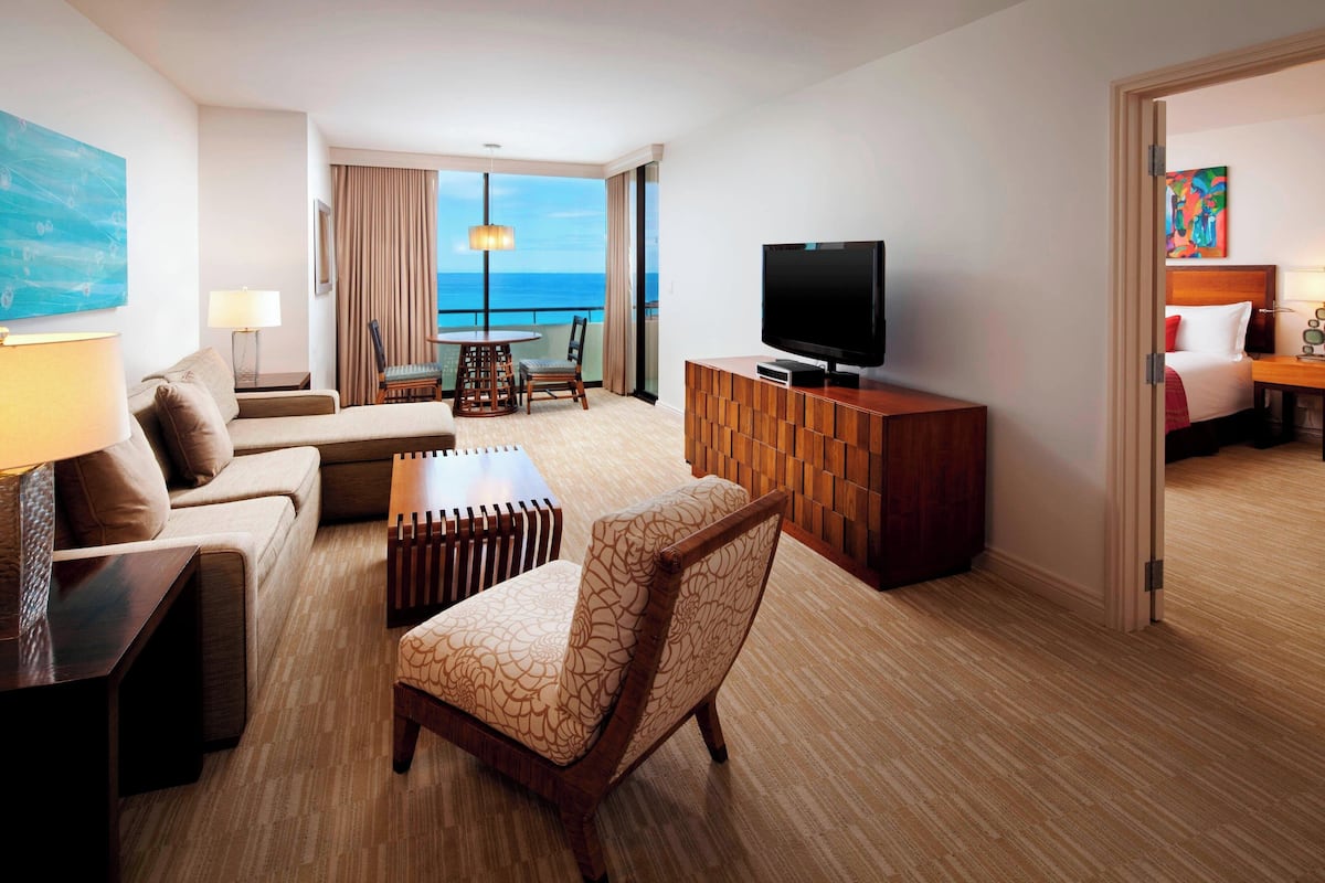A hotel room with a sofa, armchair, TV, and a view of the ocean.