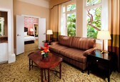A hotel room with a brown sofa, a wooden coffee table with a vase of flowers, a bedside table with a lamp, and a view of greenery through the window.