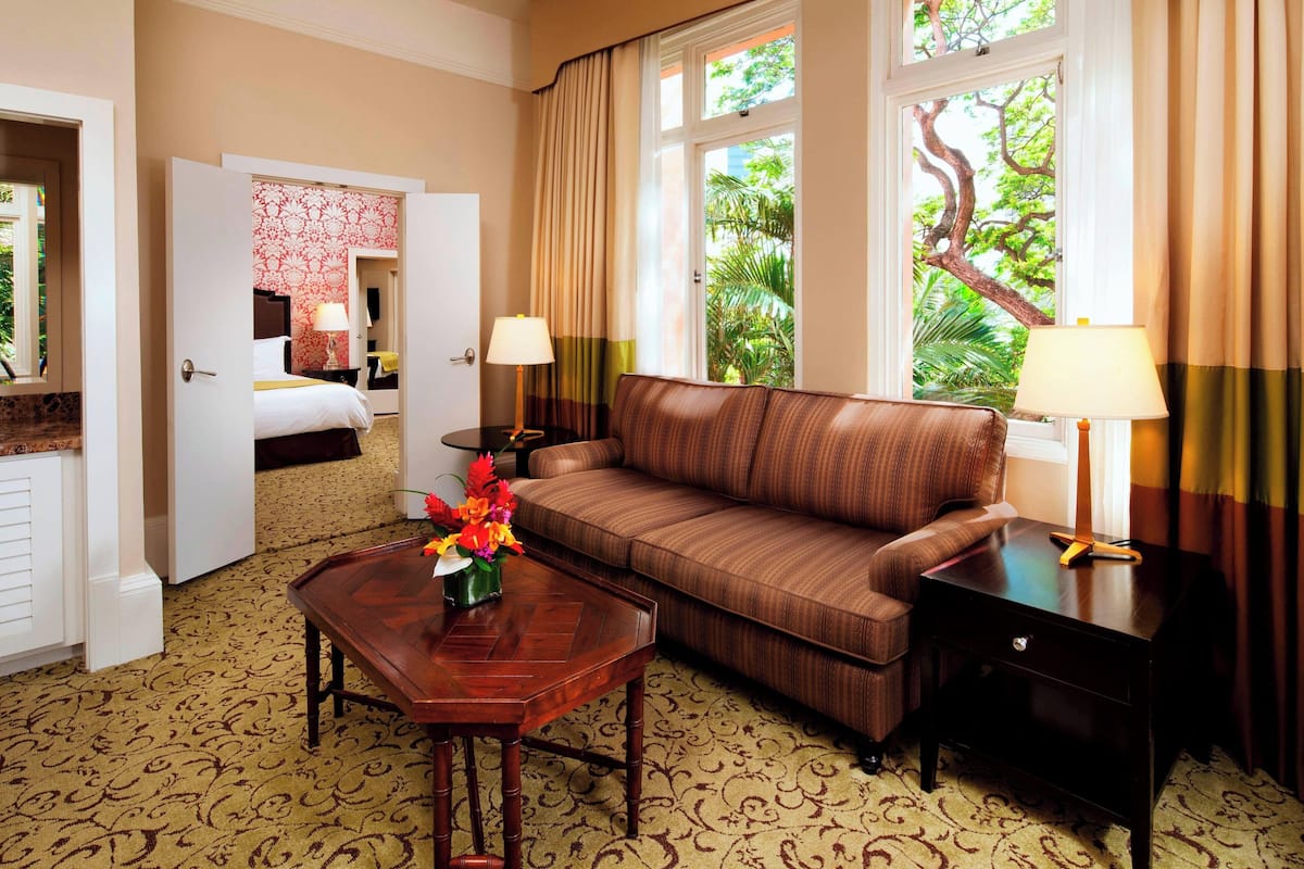 A hotel room with a brown sofa, a wooden coffee table with a vase of flowers, a bedside table with a lamp, and a view of greenery through the window.