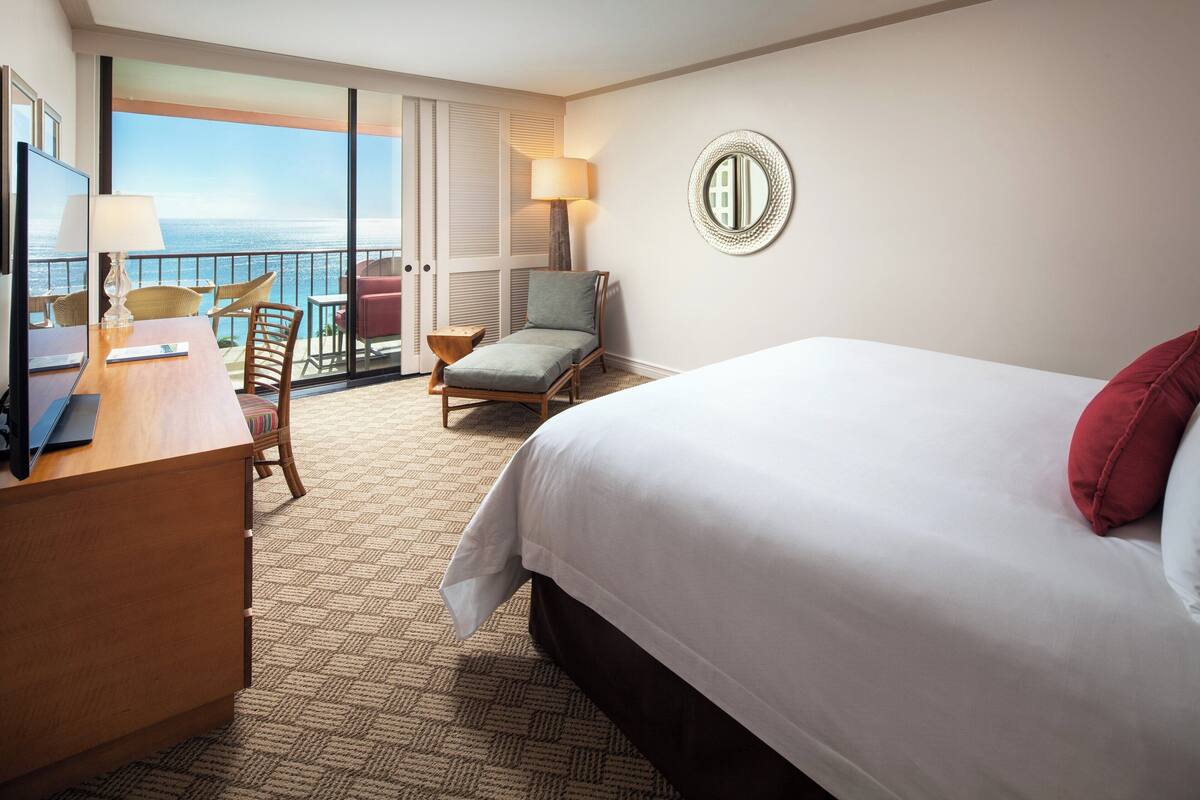 A hotel room with a bed, a desk, a chair, a lamp, and a view of the ocean.