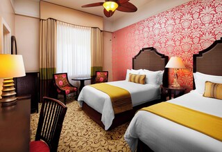 A hotel room with two beds, a desk, a chair, and a ceiling fan.