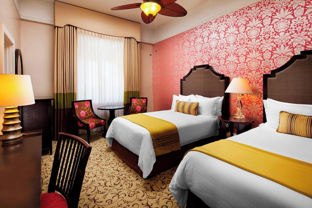 A hotel room with two beds, a desk, a chair, and a ceiling fan.