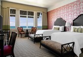 A hotel room with two beds, a desk, a chair, and a view of the ocean.