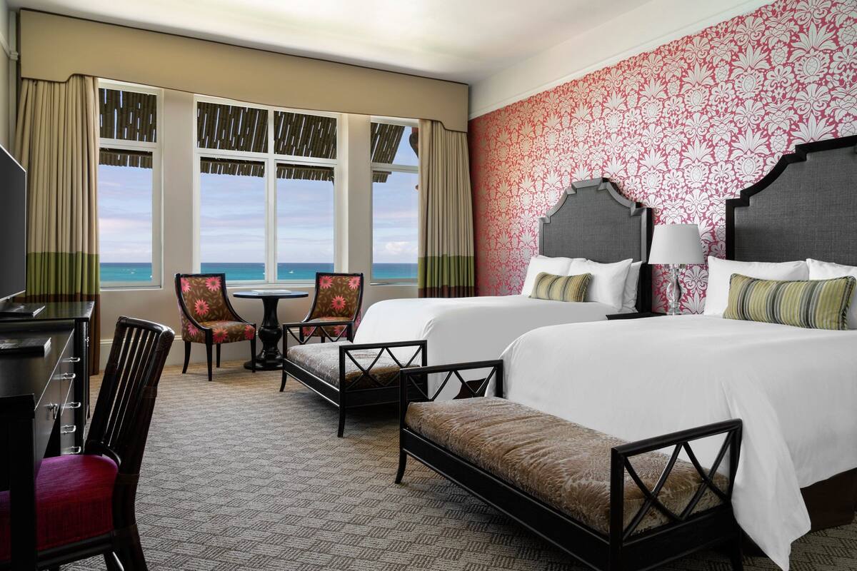 A hotel room with two beds, a desk, a chair, and a view of the ocean.