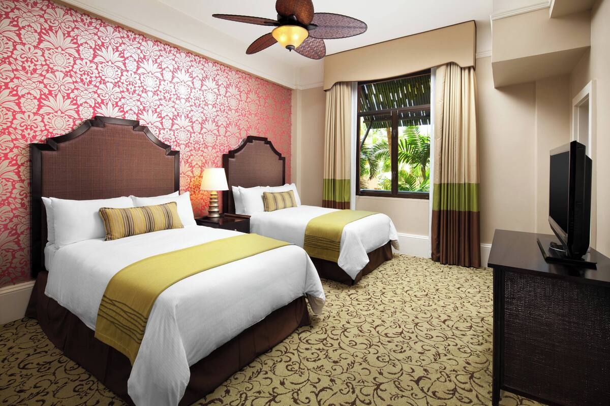 A hotel room with two beds, a ceiling fan, a television, and a window with curtains.
