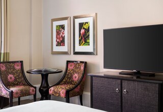 A hotel room with two floral armchairs, a small round table, a flat-screen TV, and a dark wooden cabinet.