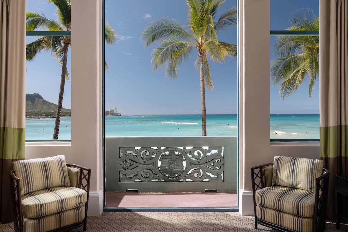 A room with a view of the ocean and palm trees, featuring two striped armchairs and a decorative screen.