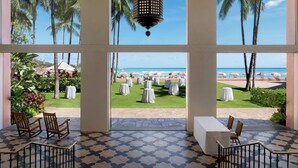 Vue depuis une chambre d’hôtel, avec de grandes fenêtres donnant sur une plage où sont disposées des tables et des chaises pour les repas.