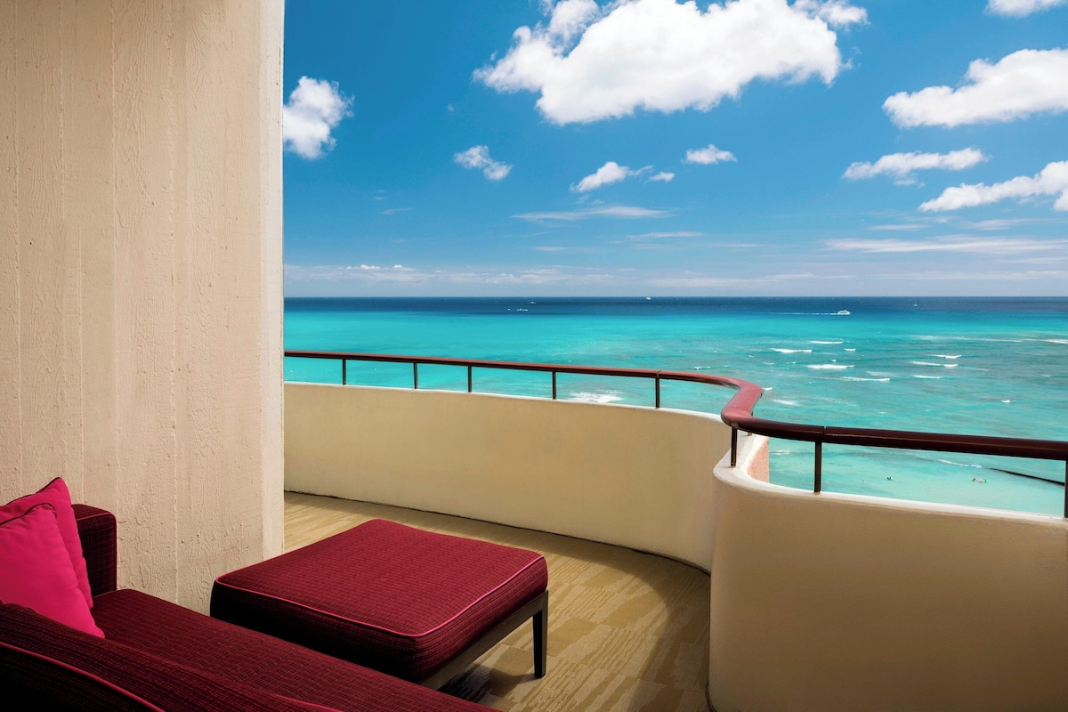 A balcony with a view of the ocean, a red cushioned chair, and a white wall.