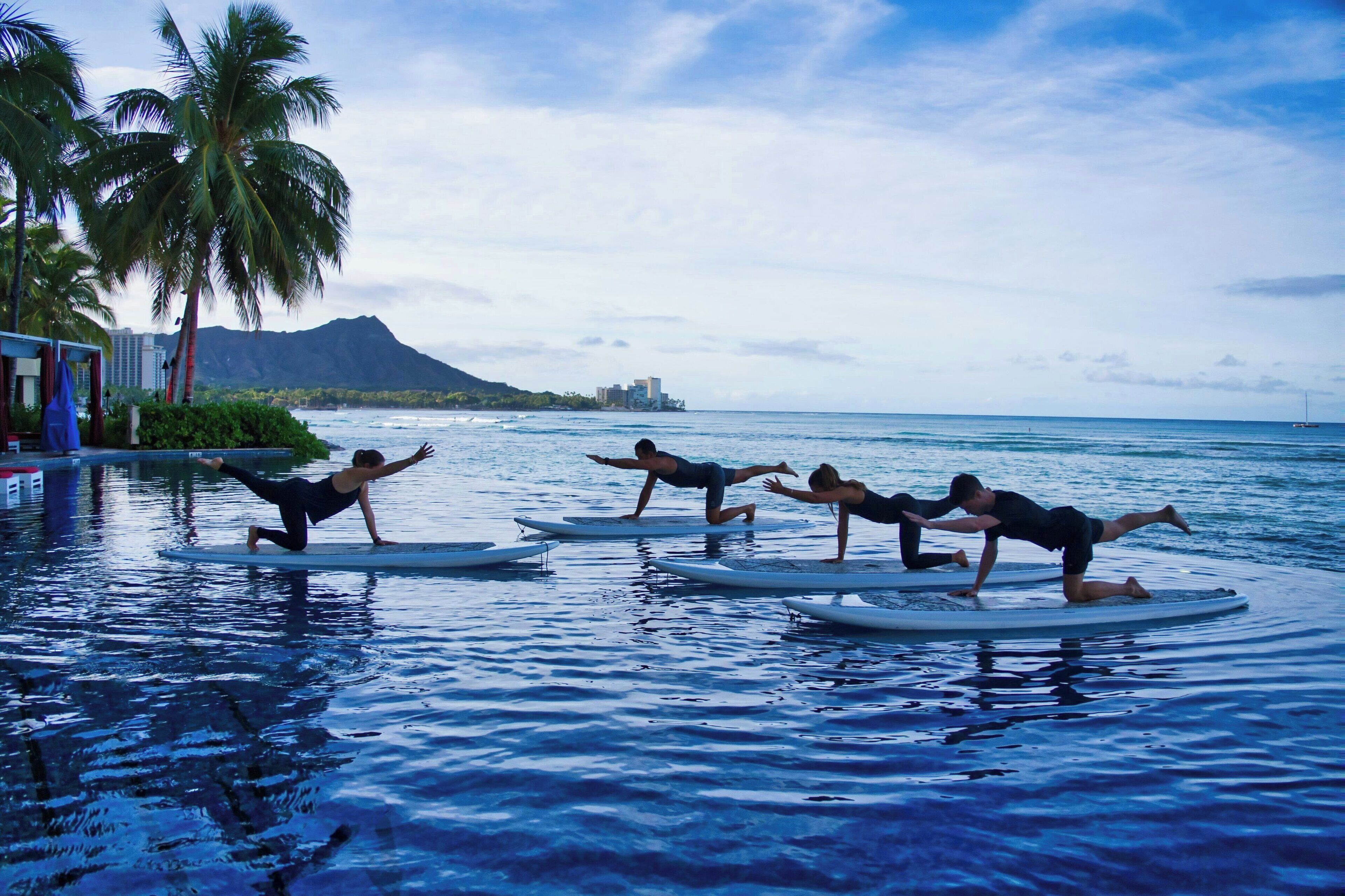Un groupe de personnes pratiquant le yoga sur des planches de surf dans une piscine, avec une vue sur une montagne et une plage en arrière-plan.