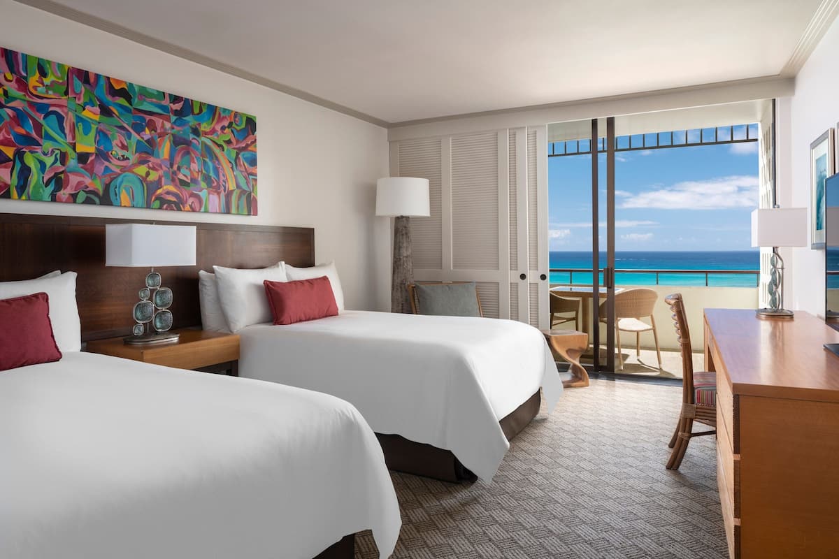 A hotel room with two beds, a desk, a chair, a large window with a view of the ocean, and a colorful abstract painting on the wall.