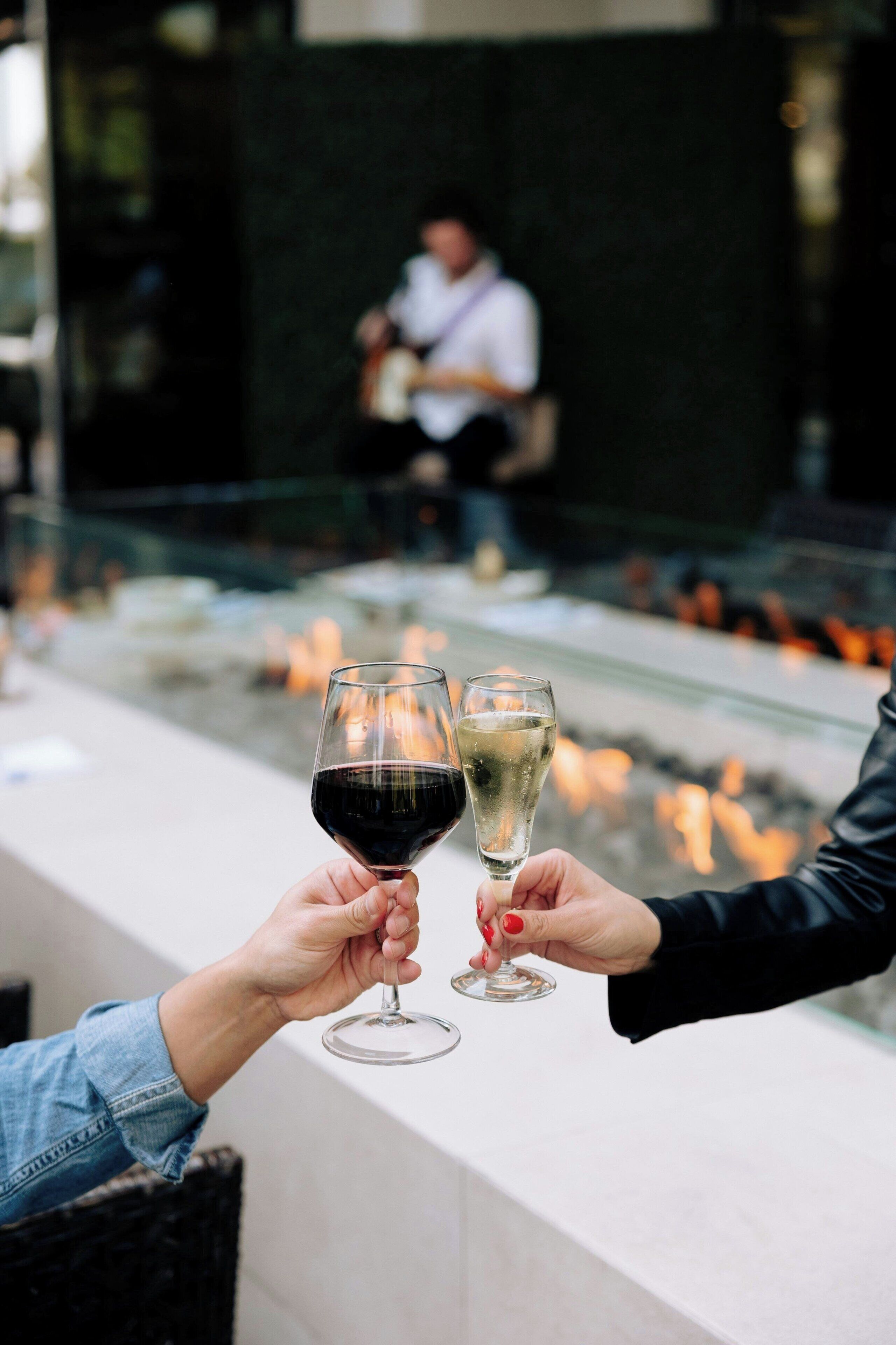 Zwei Personen stoßen mit Weingläsern an, ein Glas Rotwein und ein Glas Weißwein, mit verschwommenem Hintergrund einer Bar.