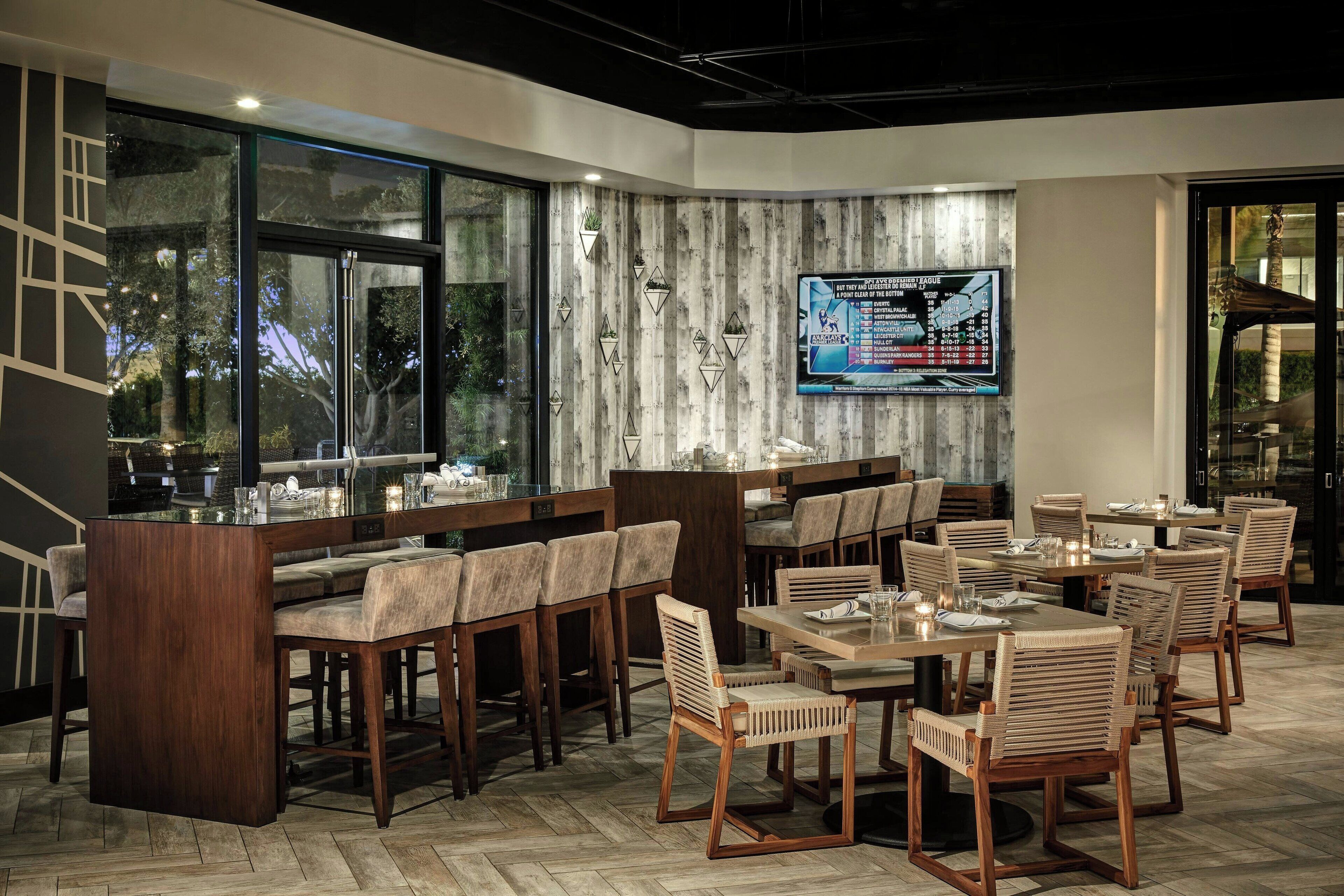 Een restaurant met een bar, houten stoelen en tafels. Er is een groot raam en een aan de muur bevestigde televisie waarop een menu wordt getoond.