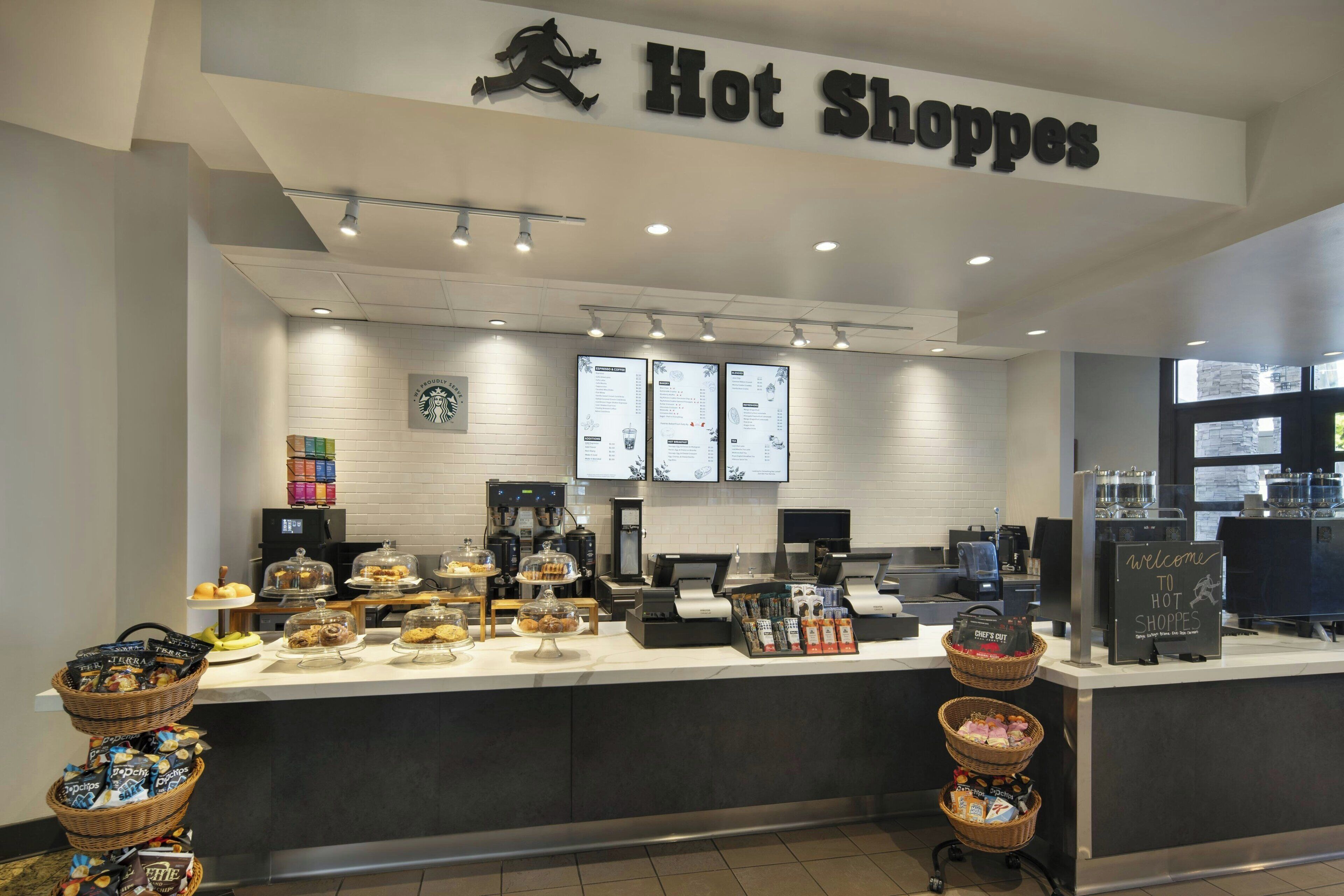 Interior d'una cafeteria amb un taulell que mostra diferents pastissos i un cartell que diu "Hot Shopspe".
