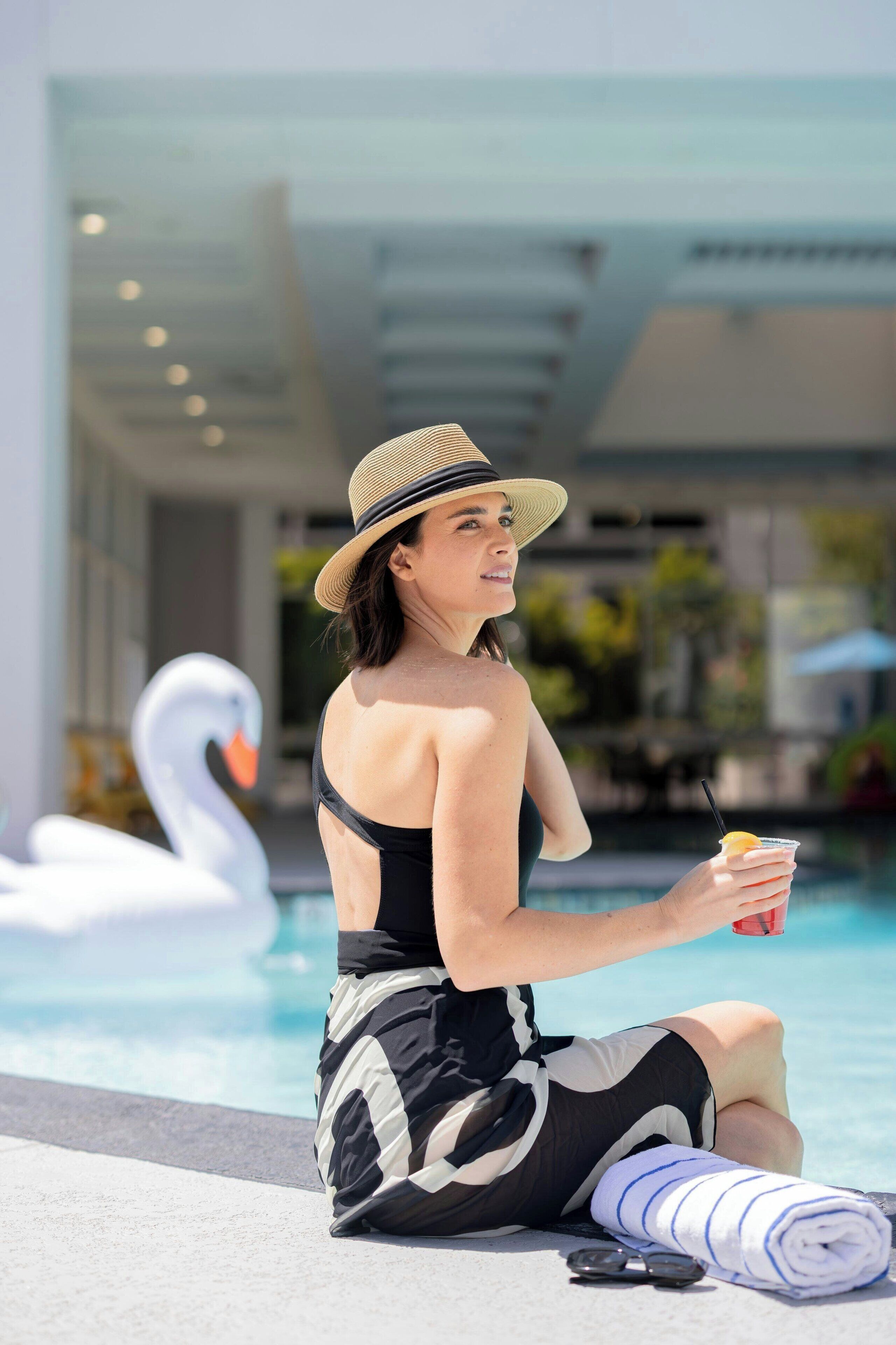 A person sitting by a pool with a swan decoration, holding a drink, and wearing a hat.