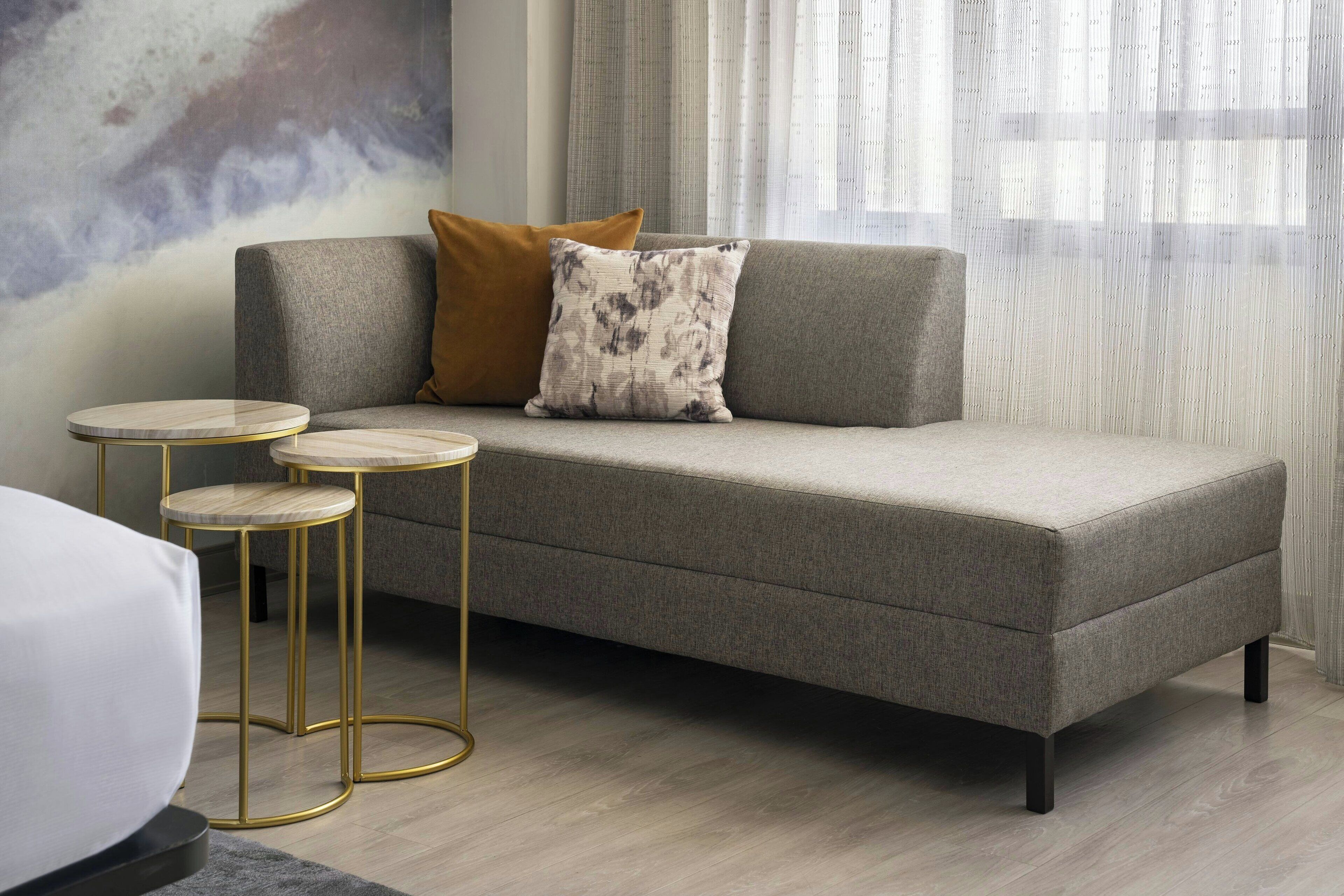 Una sala de estar moderna con un sofá gris, dos mesas redondas doradas y un amplio ventanal con cortinas translúcidas.