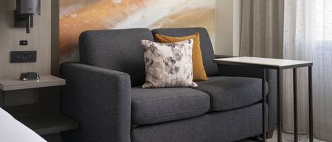 A modern living room with a grey sofa, a small side table, a wall-mounted lamp, and a large abstract painting.