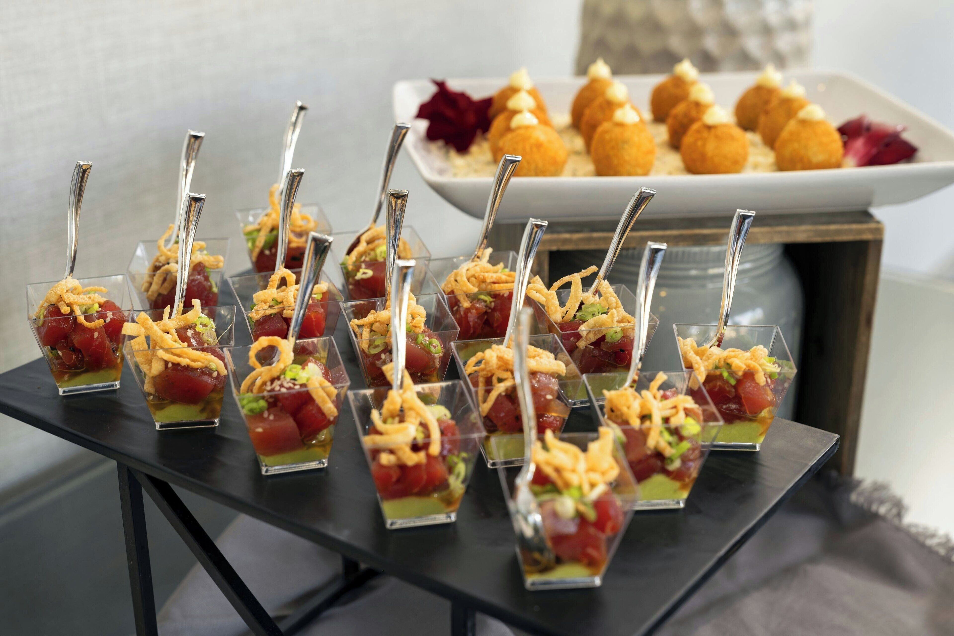 A table with clear glass cups containing a layered dessert, accompanied by a plate of round, golden-brown pastries in the background.
