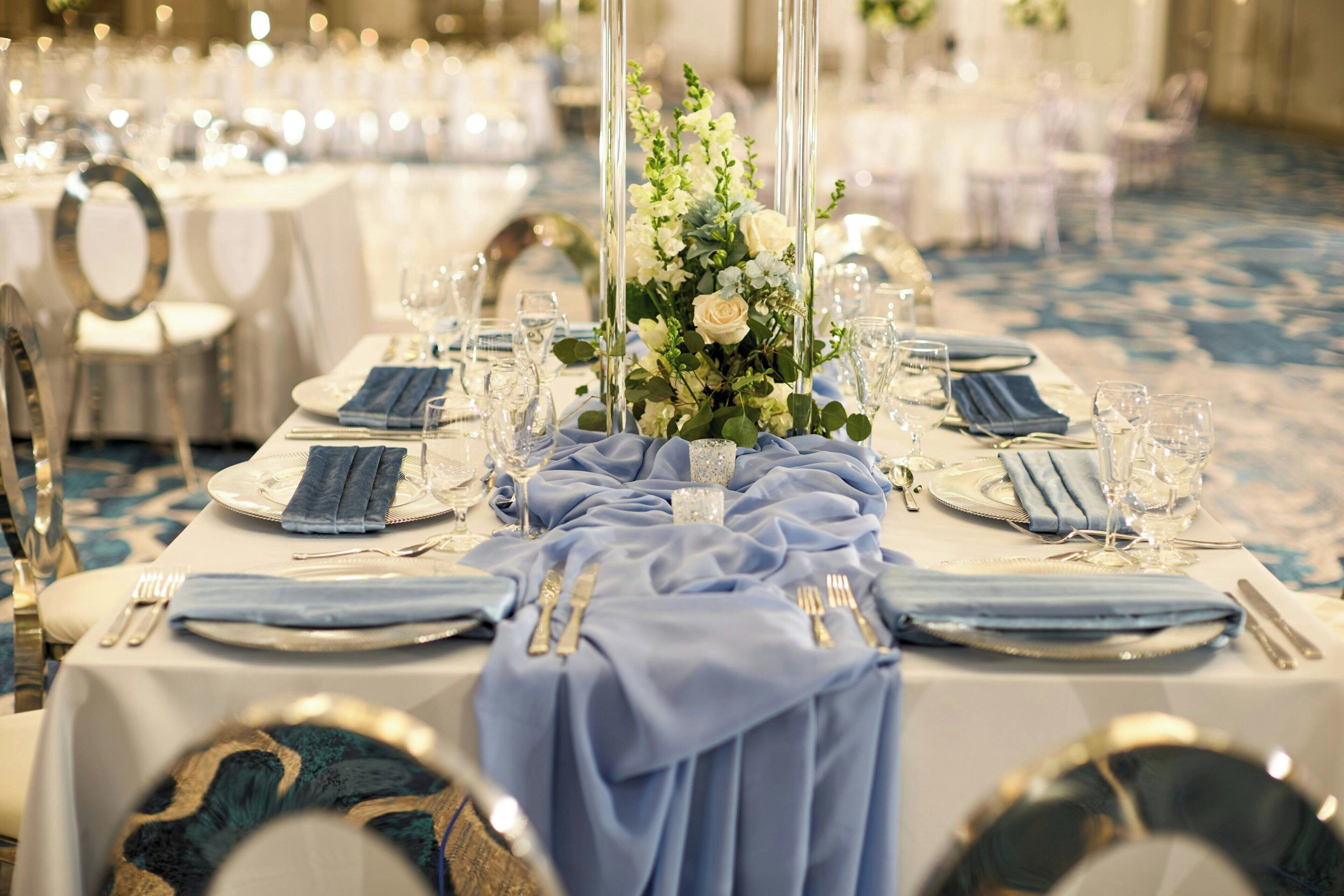 A banquet table set with blue napkins, silverware, and glassware, centered with a floral arrangement.