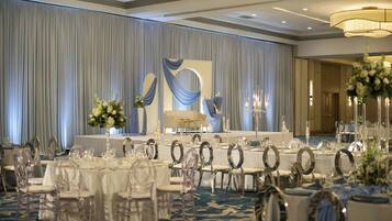 Un salón de banquetes con mesas redondas, sillas transparentes y un escenario con cortinas.