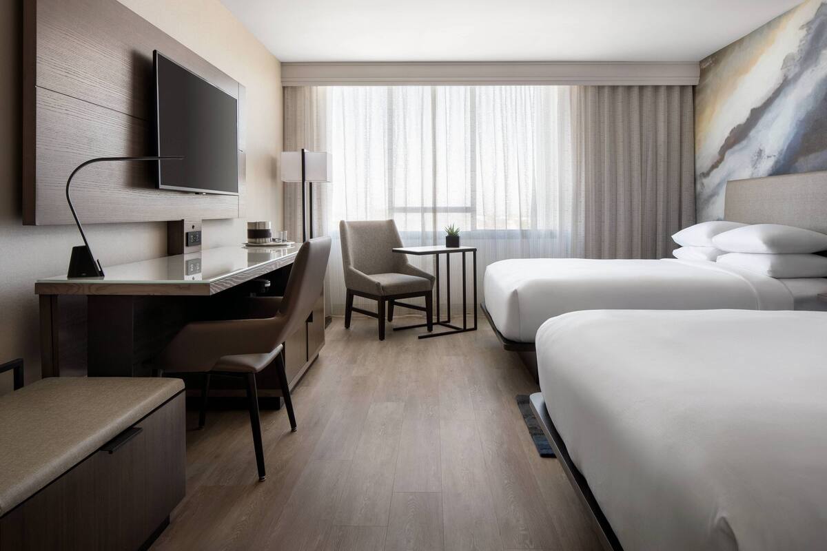 Una habitación de hotel con televisor de pantalla plana en la pared, un escritorio con silla, una mesita con lámpara, dos camas y un amplio ventanal con cortinas traslúcidas.