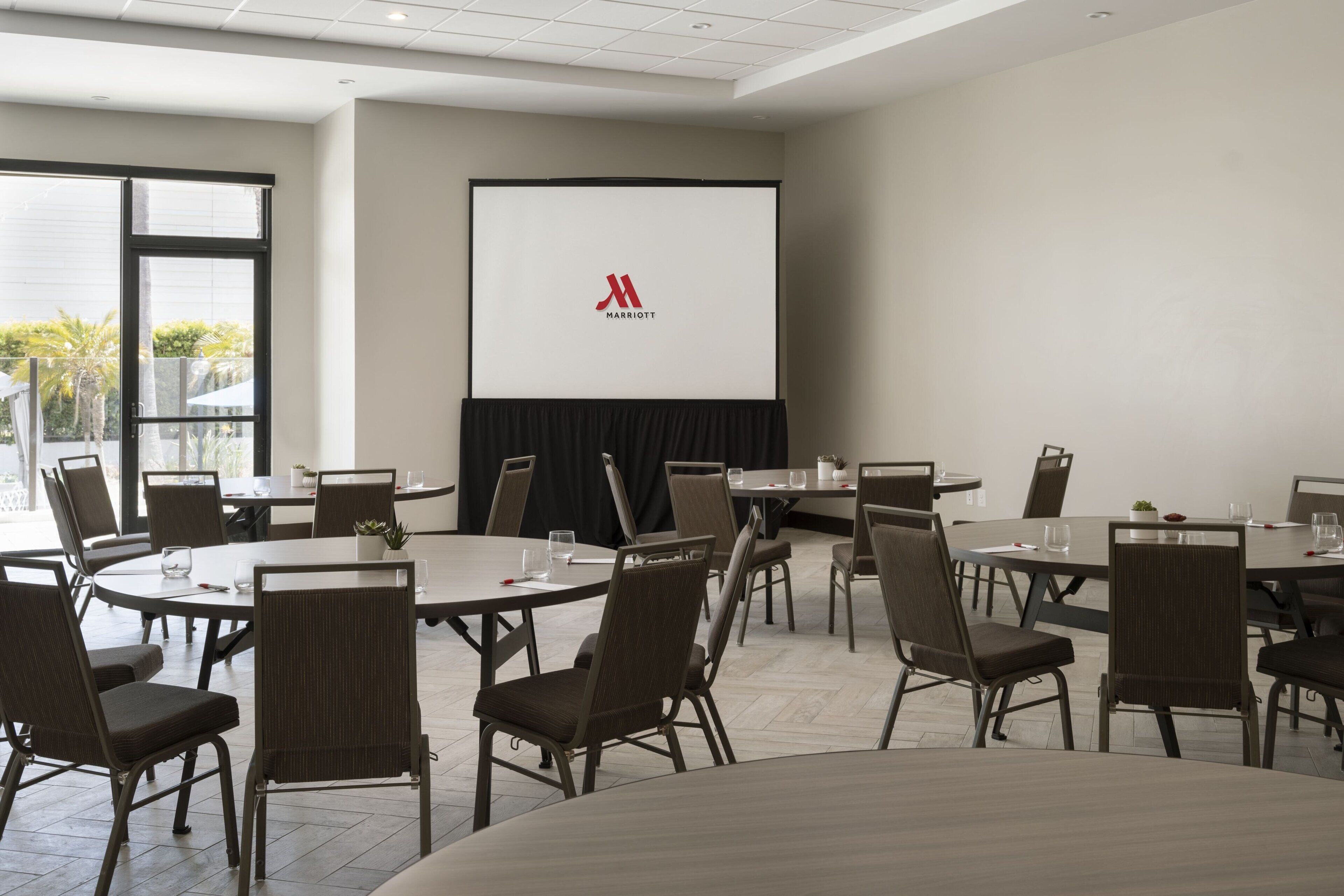 Ein Konferenzraum mit runden Tischen und Stühlen, einer Projektionsleinwand mit einem Logo und großen Fenstern mit Blick auf Palmen.