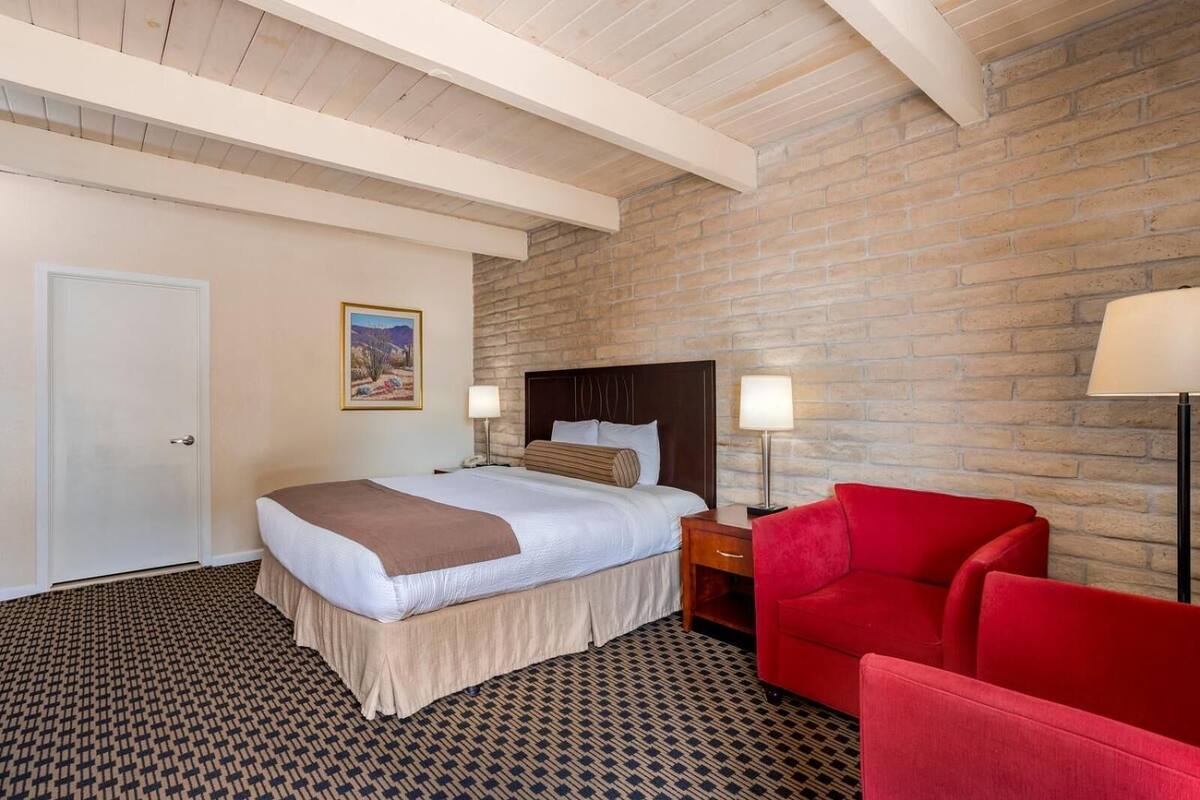 Une chambre d’hôtel avec un lit, une chaise rouge, une table de chevet, une lampe et un mur en briques.