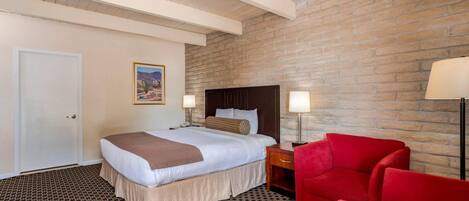 A hotel room with a bed, a red chair, a nightstand, a lamp, and a brick wall.