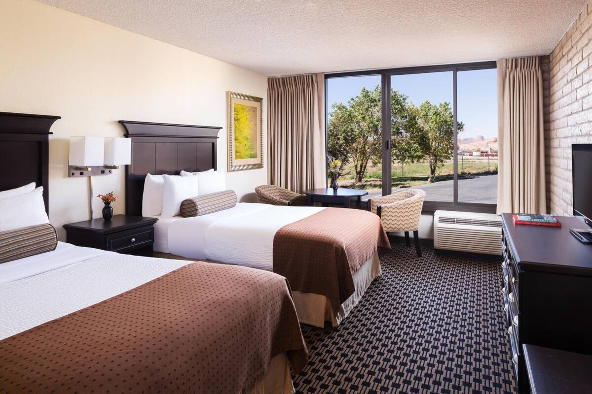 Une chambre d’hôtel avec deux lits, un bureau, une chaise, une grande fenêtre donnant sur un beau paysage et une cheminée.