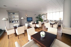 Un amplio salón con suelo de madera, mobiliario blanco, una estantería, un televisor y un ventanal grande que proporciona luz natural.