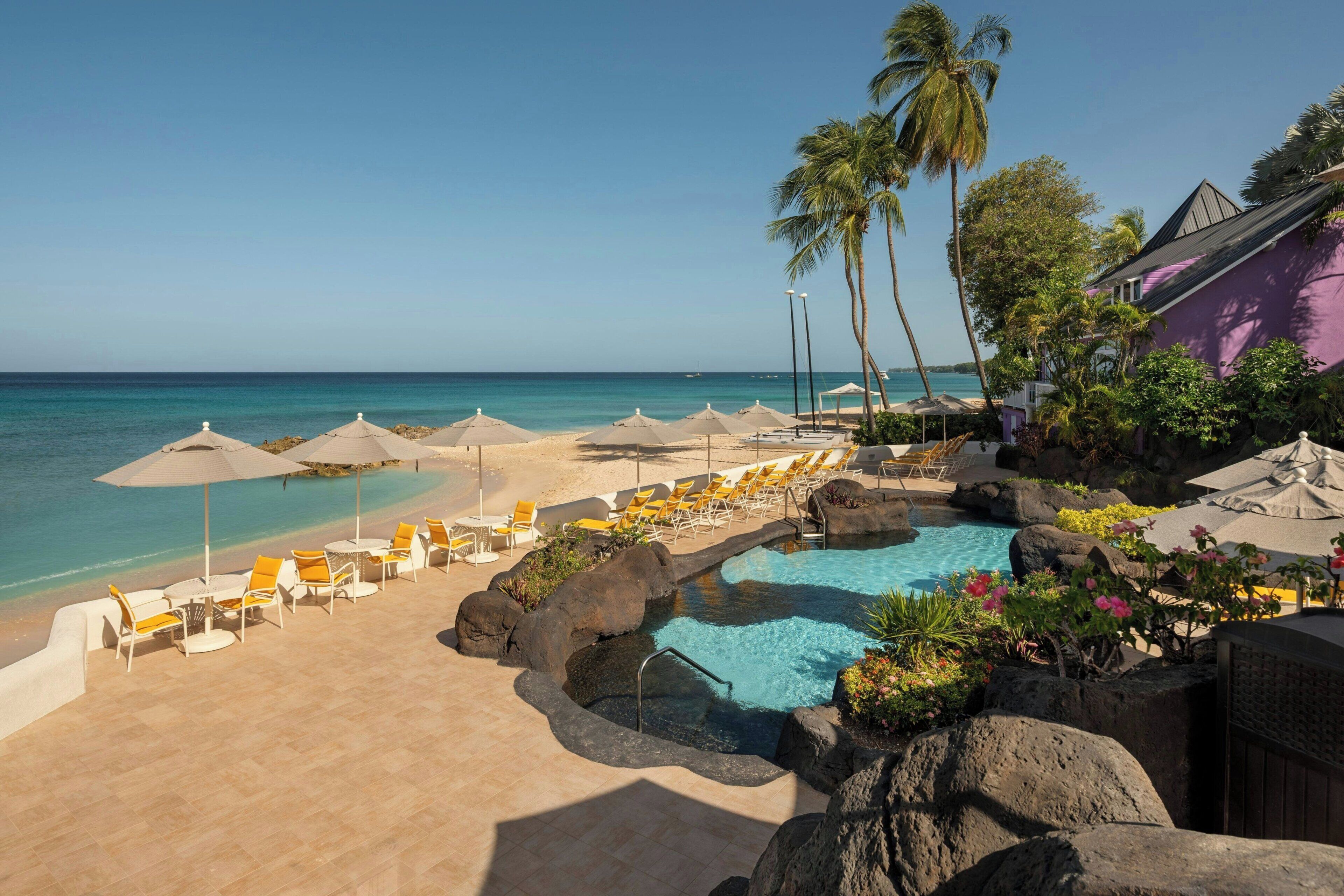 A pool area with lounge chairs and umbrellas, a clear view of the ocean, and a building with a purple facade.