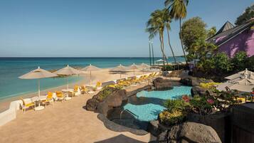 A pool area with lounge chairs and umbrellas, a clear view of the ocean, and a building with a purple facade.