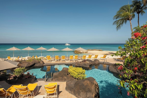 A pool with rock formations, yellow lounge chairs, and umbrellas, overlooking a beach and ocean. - Crystal Cove, A Tribute Portfolio All-Inclusive Resort (Derricks)