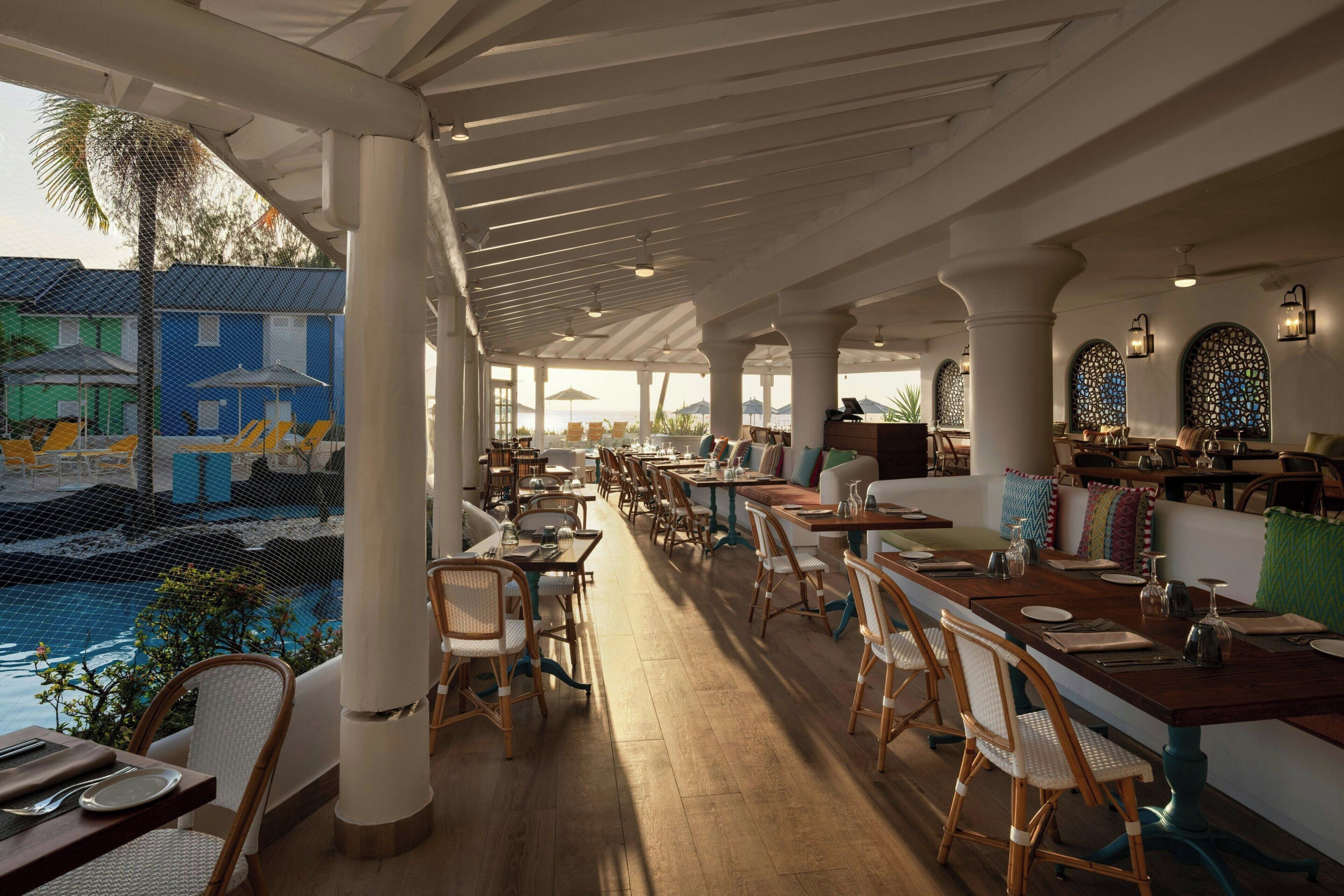 A restaurant with outdoor seating overlooking a pool, featuring a covered patio, wooden tables, and chairs.