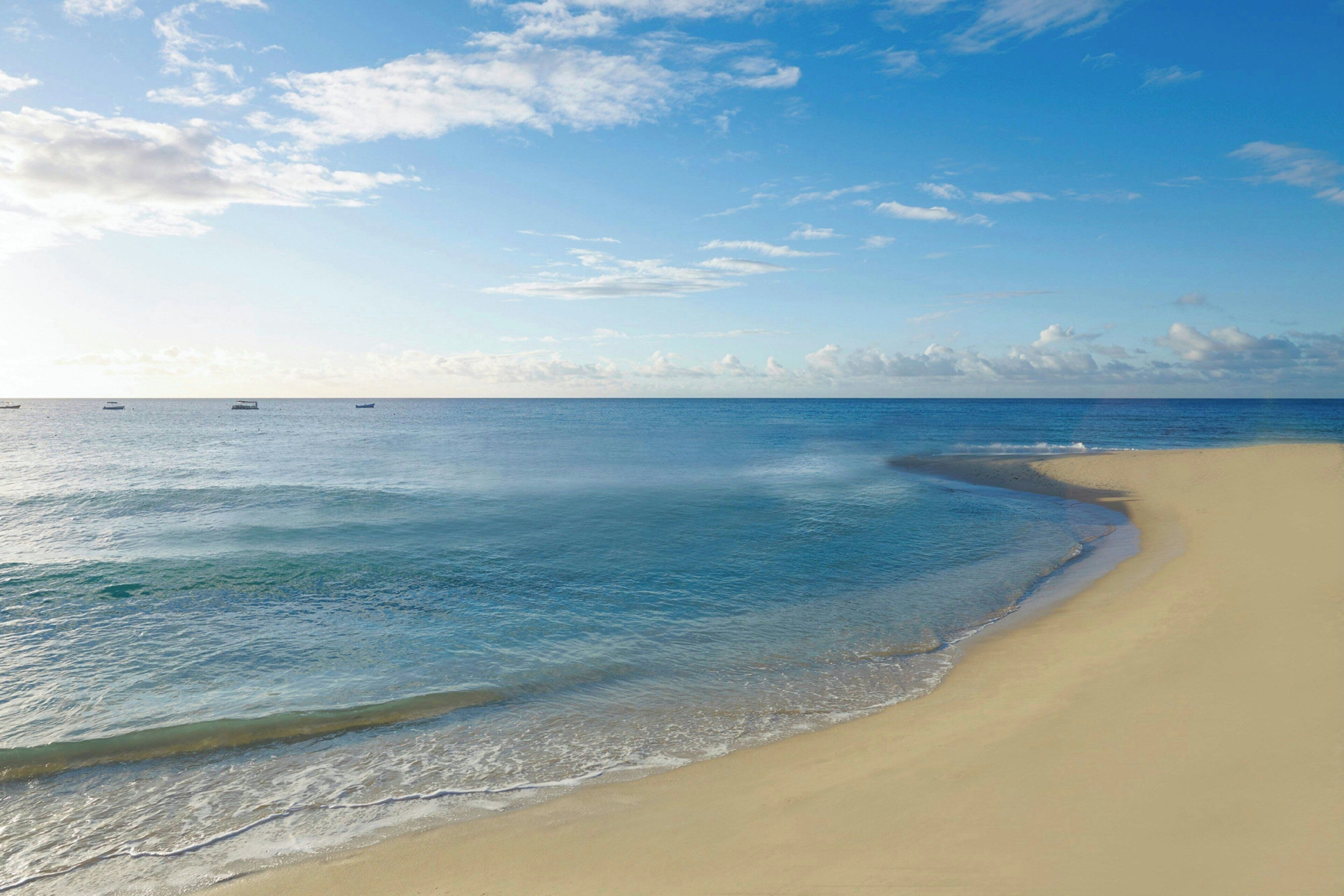 A beach with clear blue water, a sandy shore, and a few boats in the distance.