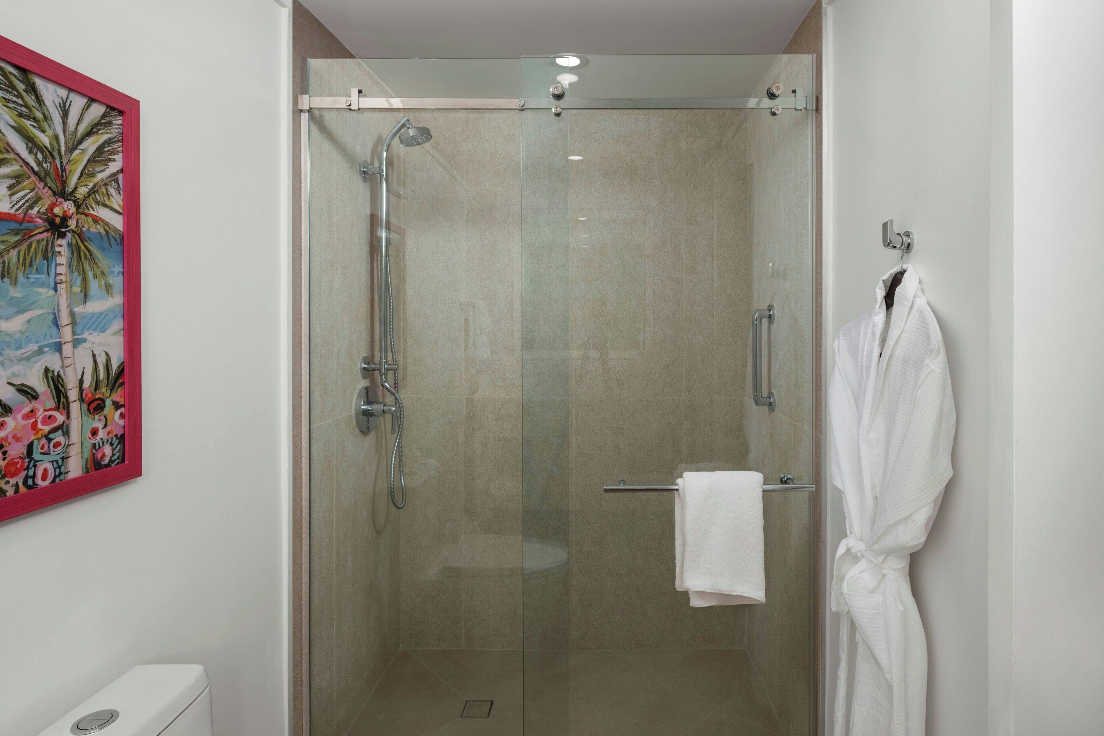 A glass-enclosed shower with a handheld showerhead, a towel rack with a white towel, and a painting of a palm tree on the wall.