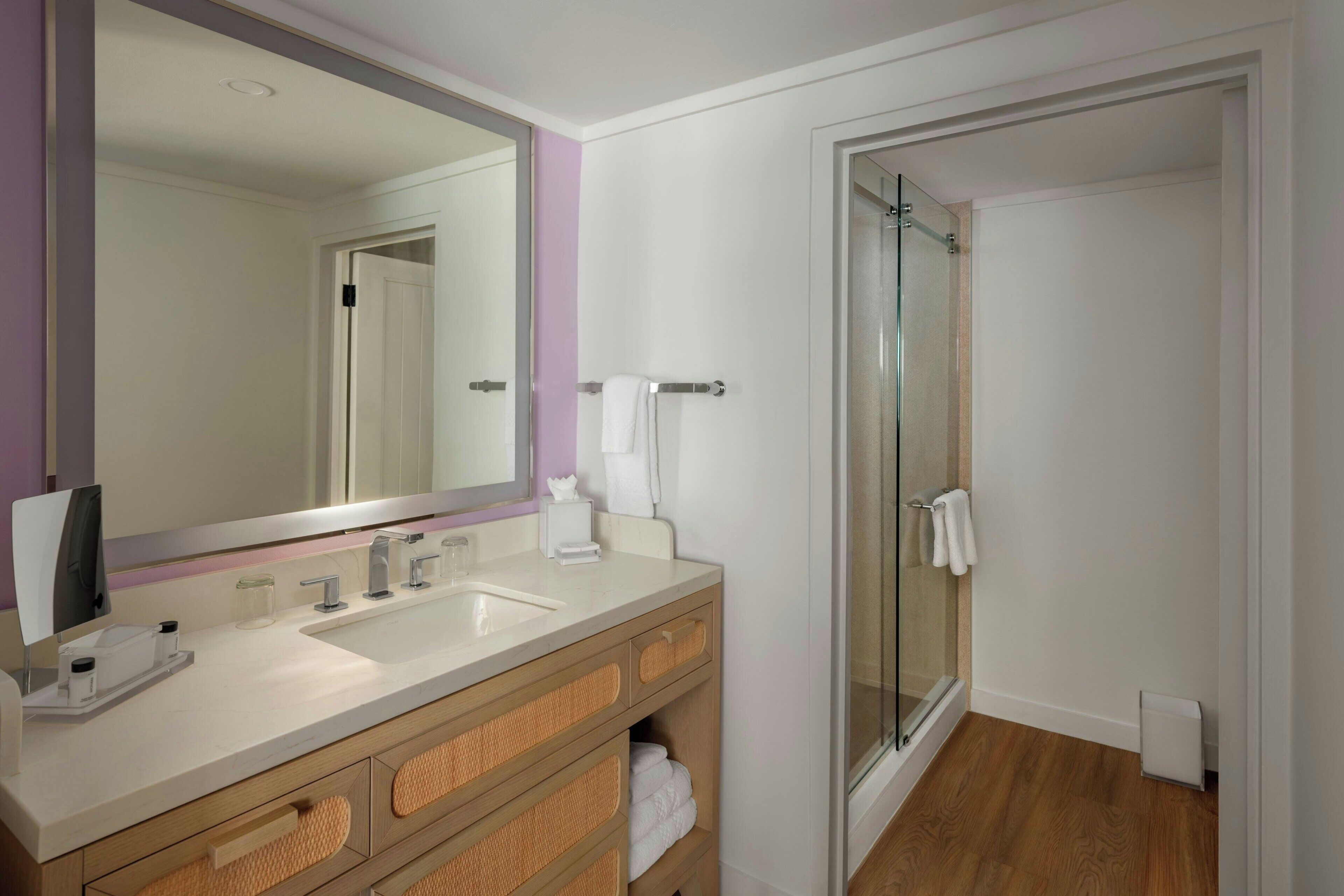 Une salle de bain dotée d'un grand miroir, de deux lavabos, d'un meuble-lavabo avec des rangements en osier et d'une cabine de douche en verre.